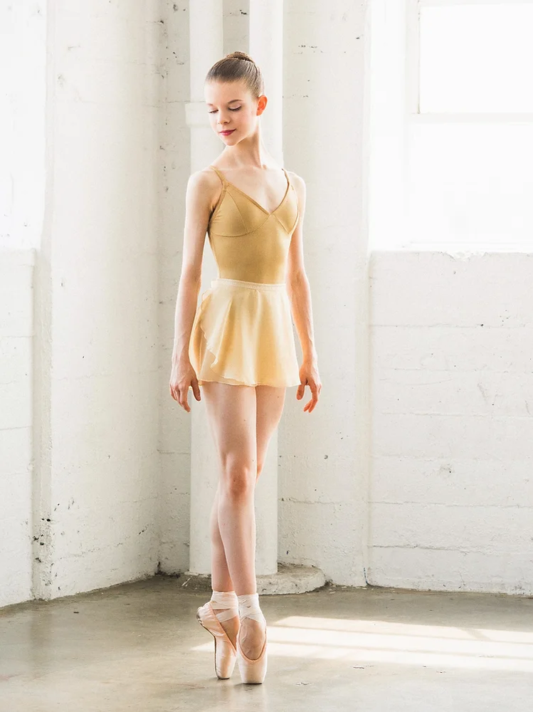 
                      
                        A young dancer stands en pointe, her hair is pulled tight into a ballet bun. She is gazing off to the side and downward. She is wearing a butter colored leotard and short wrap skirt. The background is a white stone wall and industrial pipes.
                      
                    