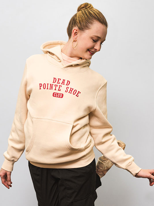 A female with a blond bun looks off to her left, back leg raised behind her. She wears black warm-up pants, pointe shoes, and a cream hoodie with red text, "Dead Pointe Shoe Club."