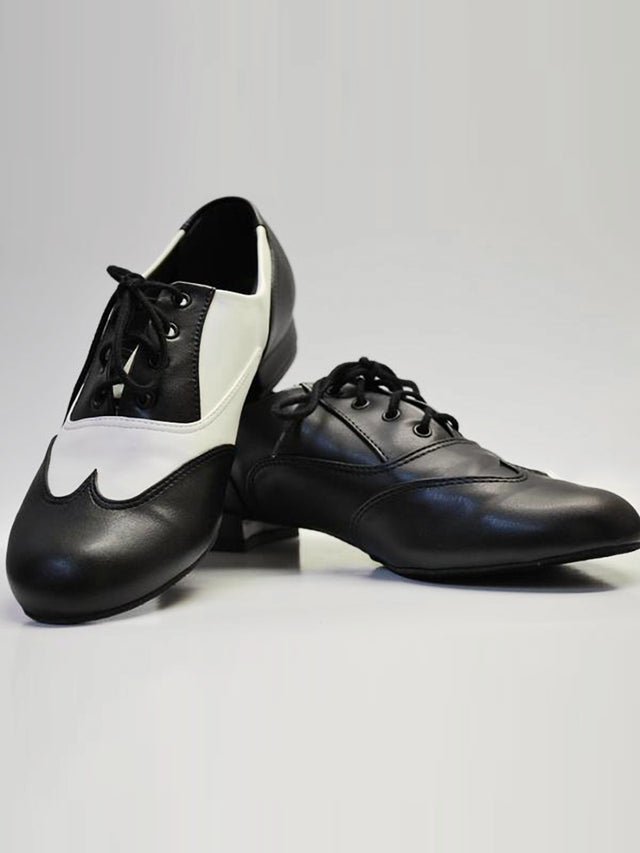 A pair of shoes sit next to each other, the heel of the left propped on on top of the right shoe. The left showcases the black and white design, the right the all black design. Both feature oxford stitching and lace-up closure with a small standard heel. Grey background.