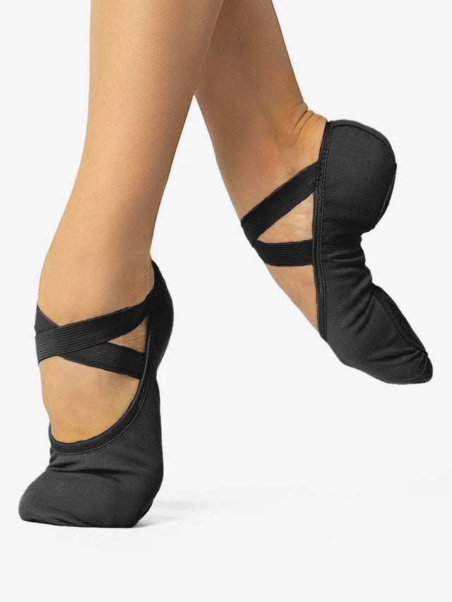 A close-up of a person's legs, wearing black, canvas ballet shoes with a criss-cross elastic design and no drawstring. The front leg is in releve and the back leg lifted and pointed to show the flexible design and contour, against a plain background.