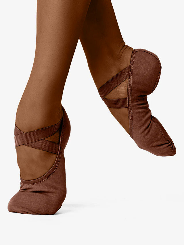 A close-up of a person's legs, wearing mocha skin-tone, canvas ballet shoes with a criss-cross elastic design and no drawstring. The front leg is in releve and the back leg lifted and pointed to show the flexible design and contour, against a plain background.