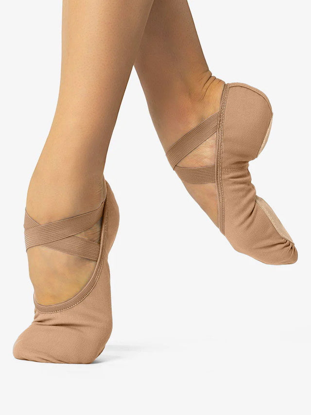 A close-up of a person's legs, wearing suntan skintone, canvas ballet shoes with a criss-cross elastic design and no drawstring. The front leg is in releve and the back leg lifted and pointed to show the split-sole design and contour, against a plain background.