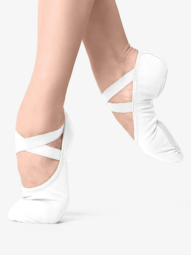 A close-up of a person's legs, wearing white, canvas ballet shoes with a criss-cross elastic design and no drawstring. The front leg is in releve and the back leg lifted and pointed to show the split-sole design and contour, against a plain background.