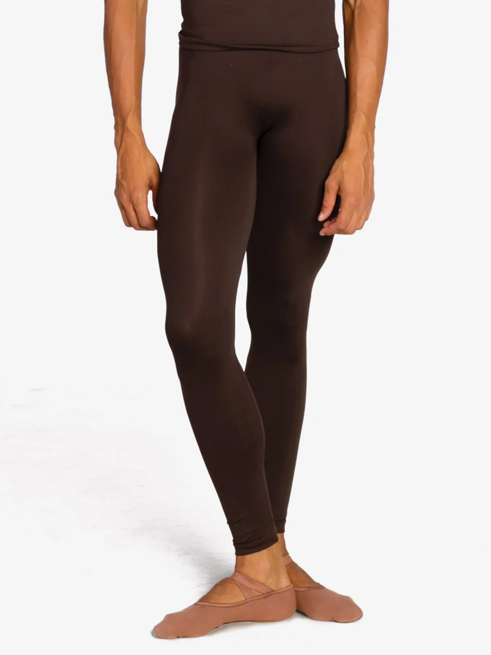 A close-up of a male dancer from the waist down wearing coffee colored tight leggings and nude canvas ballet shoes against a plain background.