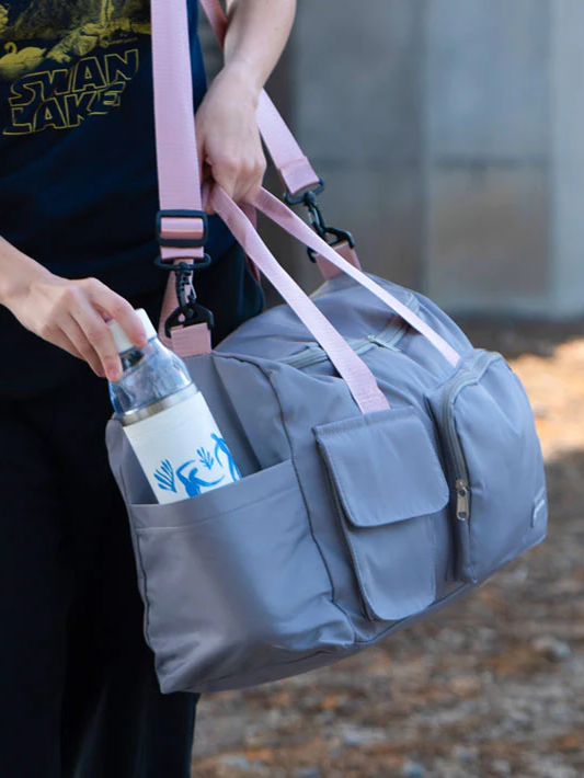 
                      
                        A close-up of a person carrying a grey duffle bag with pink straps, as they put a water bottle in one of its many pockets.
                      
                    