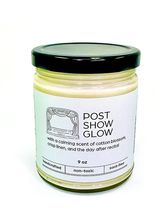 A white candle with a black lid, with a label and title "Post Show Glow" in cotton blossom and crisp linen scent.