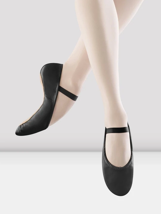 A close-up of a person's legs wearing black, leather ballet shoes with a single elastic strap across the top of the foot. The feet are pointing down as if mid-air, the back leg bent to show the flexibility of the shoe and pointe of the foot.